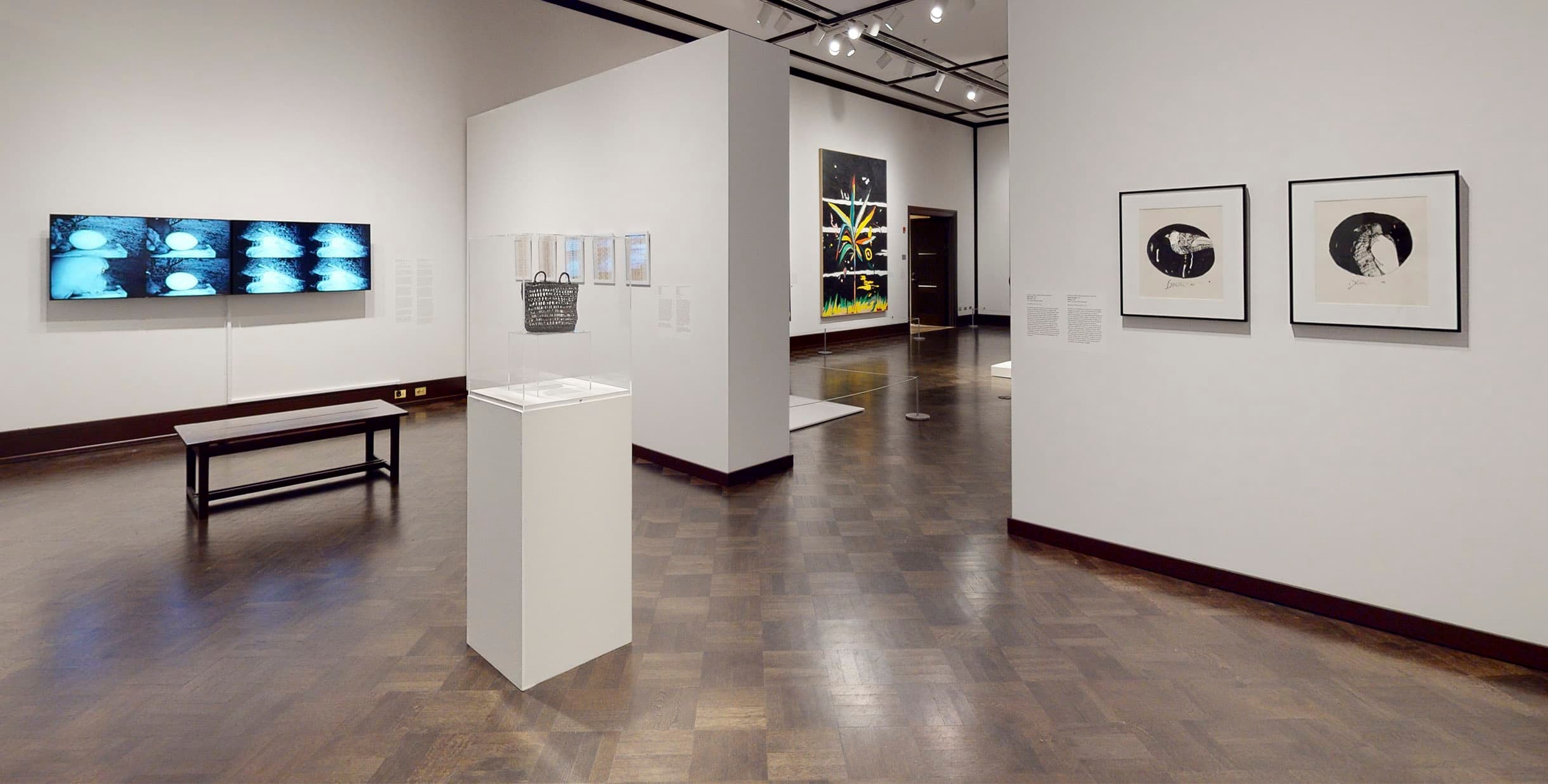 Focus Central gallery White walled gallery with several framed artworks and a video screen. There is a bench at the far end and a pedestal with an artwork in the center of the room. Through a doorway a glimpse of another gallery is visible.